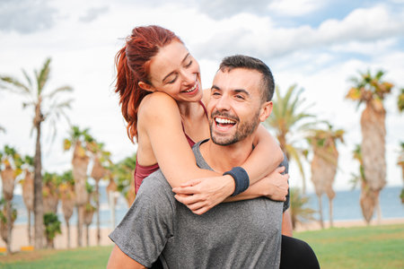 Man smiling and carrying his girlfriend piggyback after their cardio workout. Young athletic couple enjoying together, hugging and looking at eachの写真素材