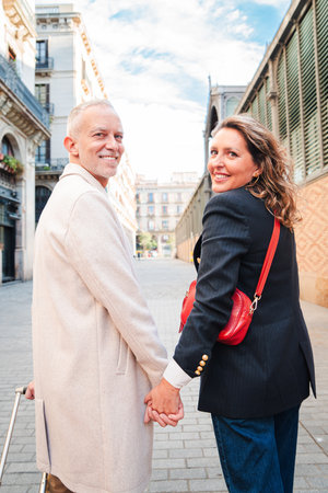 Back view of a middle aged married couple holding hands while smiling and looking at camera. Mature husband and wife having a good time on a romantic trip in Europe. Happy mid adult mates enjoying. High quality photoの写真素材