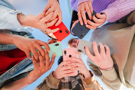 Vertical low angle view of a group of unrecognizable multiracial young friends using their cellphones on a circle. Team of teenagers enjoying andの写真素材