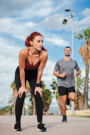 Vertical. A tired sports woman resting after the effort made during cardio training, while a athletic young man runs. An exhausted female after aの写真素材
