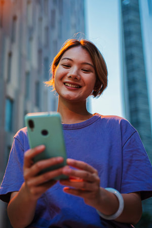 Nighttime: Vertical portrait of a 20 years Asian young girl engrossed in her smartphone to browse internet on a city street at night, capturing aの写真素材