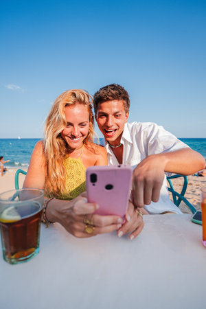 Vertical. Young couple using a smart phone to browse on internet sitting at a beach bar during their summer vacation on the coast. Boyfriend andの写真素材