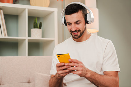 Smiling man wearing headphones using a yellow smartphone at home, likely listening to music or texting, sitting on a couch in a relaxed indoorの写真素材