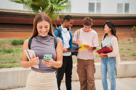 On foreground one blonde teenage female student using her smartphone at university campus, at background a group of students talking together. Cherfulの写真素材