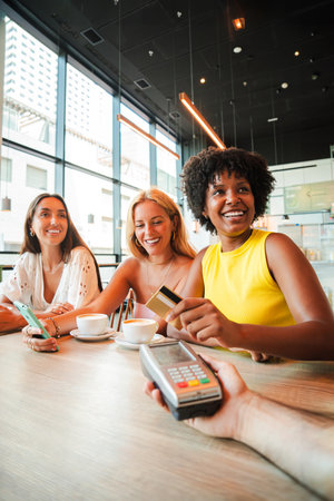 Vertical. Young African American woman is settling her bill with a contactless creditcard at a restaurant. She smiles as she hands over her creditの写真素材