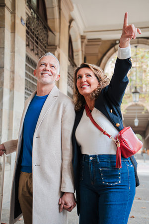 Vertical Mature couple, pointing and walking with a suitcase, enjoying their vacation. Middle-aged pair is having fun sightseeing in a European cityの写真素材