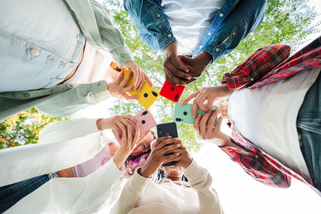 Low angle view of group of friends standing in circle, all using cellphones outdoors. Vibrant smartphone colors contrast the sky, showing modernの写真素材