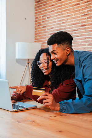 Vertical. Smiling African American couple using a laptop and credit card to make an online purchase, enjoying convenient shopping at home in a cozyの写真素材