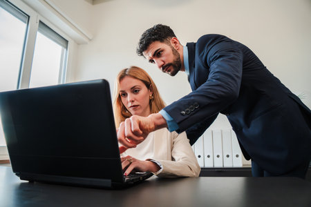 Business colleagues collaborating in an office environment, with a male professional pointing at a laptop screen while assisting a female coworkerの写真素材