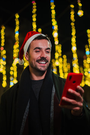 Vertical. Smiling young man wearing a Christmas hat using his cellphone at night, surrounded by festive lights, enjoying the holiday season with joyの写真素材