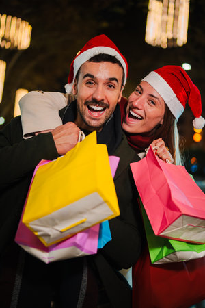 Vertical Cheerful young couple wearing Christmas hats and holding colorful shopping bags, enjoying festive holiday shopping at night with glowingの写真素材