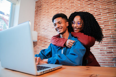 Happy African American couple enjoying time together at home, using a laptop on a cozy desk, embracing and smiling in a warm, relaxed environmentの写真素材