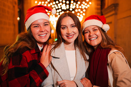 Group of cheerful young women wearing Santa hats and smiling at the camera, standing outdoors at night with festive Christmas lights glowing in theの写真素材