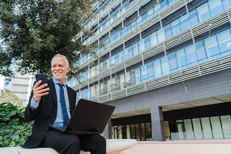Copy Space. Confident businessman multitasking with a cellphone and laptop outdoors, showcasing productivity and modern work in a professional settingの写真素材