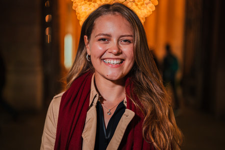 Smiling young woman with long blonde hair and a red scarf, standing at night under festive lights, looking directly at the camera with a happyの写真素材