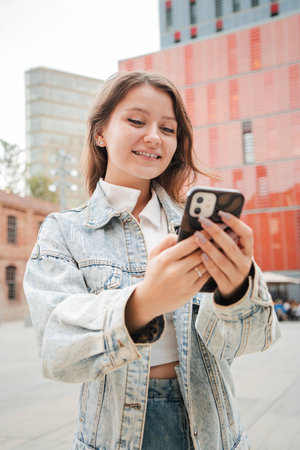 Vertical. Teenager girl smiling while using a cellphone outdoors, wearing a denim jacket, standing in front of red buildings and enjoying a casualの写真素材