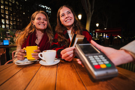 Two young women smiling while paying the coffee shop bill with a credit card at night, sitting at a wooden table outdoors with hot drinks and enjoyingの写真素材