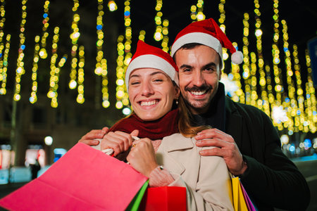 Smiling couple wearing Santa hats holding shopping bags during festive Christmas night, enjoying holiday spirit with lights, showcasing happiness andの写真素材
