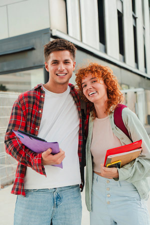 Vertical. Two cheerful classmates posing together outdoors, smiling warmly at the camera while holding books and backpacks, showcasing a friendly andの写真素材