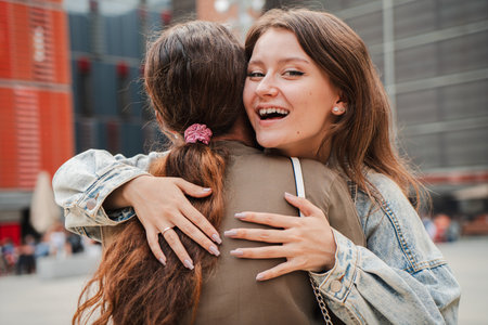 Young woman smiling warmly while hugging a friend outdoors in an urban area, expressing joy,...