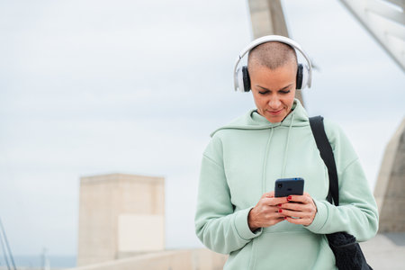 A mid adult woman enjoying music on her smartphone while wearing headphones and a stylish hoodie,...