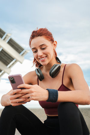 Person in athletic wear enjoying smartphone while sitting outdoors with headphones, engaged in mobile phone applicationの写真素材
