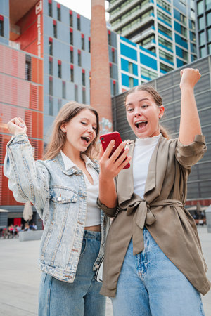 Two women celebrating their victory while using an application on their cellphones, showing excitement and joy after winning, highlighting theの写真素材