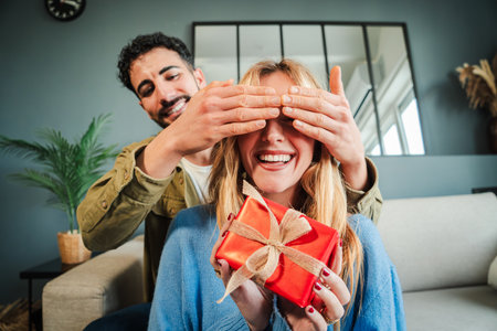 Surprising Moment of Joy: Man Playfully Covering Womans Eyes While Handing Her a Beautifully Wrapped Gift in a Cozy Living Room Filled with Delightedの写真素材