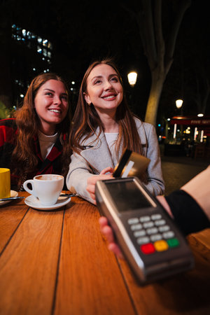 Two Friends Are Happily Engaged in a Payment Process with a Credit Card While Enjoying Their Coffee Together. Their Smiles Reflect the Delight of theの写真素材