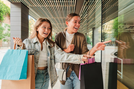 Two Happy Friends Are Enjoying A Joyful Day Of Shopping Together. They Are Laughing And Pointing Excitedly At A Storefront Display While Holdingの写真素材