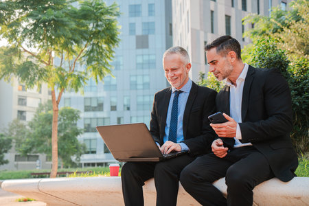 Two confident businessmen engaged in a productive discussion while using a laptop outdoors. Their professional attire reflects their dedication toの写真素材