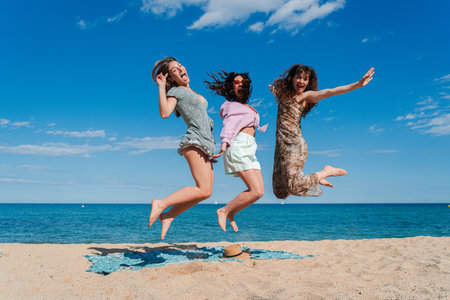 Joyful moments of three carefree young women jumping together on a sunny beach with a stunning ocean view and a clear blue sky, celebrating friendshipの写真素材