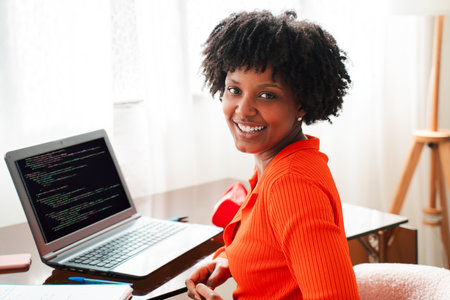 Smiling young woman sitting at her desk while coding on her laptop with programming text displayed,...
