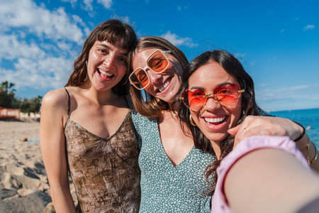 Three cheerful friends enjoying a sunny beach day while taking a selfie with smiles and laughter under the clear blue sky, capturing the essence ofの写真素材