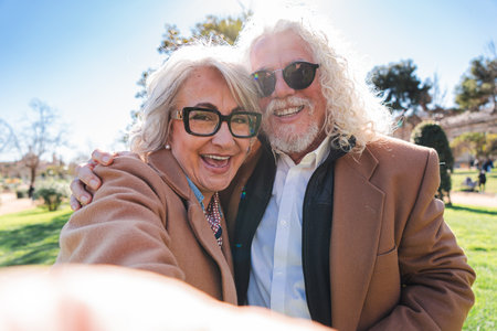 Joyful elderly couple enjoying a cheerful moment while taking a selfie in a sunny park, showcasing...