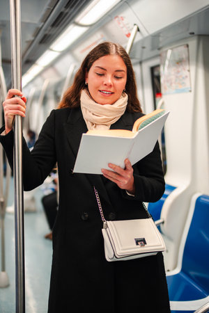 Engaged woman reading a fascinating book while standing in a subway train, enjoying her time as a passenger during the daily commute amidst fellowの写真素材