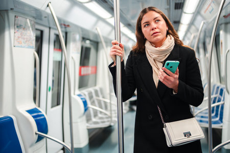 Thoughtful young woman standing in a subway train while using her smartphone and browsing the internet on her mobile phone during her daily commute inの写真素材