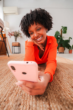 Cheerful young woman joyfully using her smartphone while lying on a cozy rug, engaging with captivating content and enjoying her leisure time in aの写真素材
