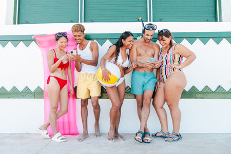 Friends enjoying a sunny day at the beach while engaging with their smartphones and sharing laughterの写真素材