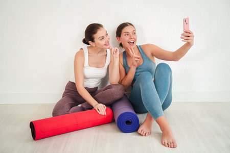 Two energetic young women joyfully taking a playful selfie together while sitting on yoga mats and enjoying a moment of friendship and fun in a brightの写真素材