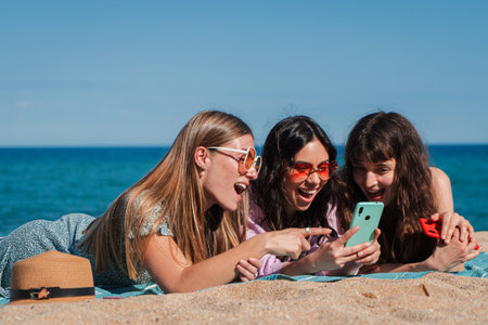 Three excited young women enjoying a fun moment together while using a smartphone on the beachの写真素材