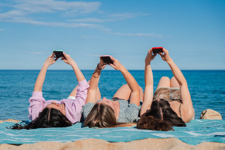 Three joyful women lying on the beach while using their smartphones to capture fun moments togetherの写真素材