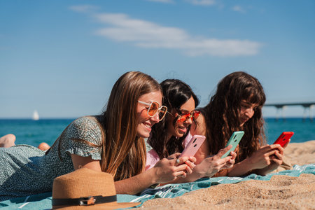 Three cheerful young women enjoying their time at the beach while using their smartphones and laughingの写真素材