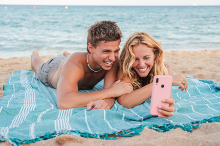 Joyful couple enjoying a fun selfie moment together while lying on a beach towel at the sandy shoreの写真素材