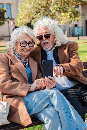 Joyful elderly couple sharing a delightful moment while using a smartphone to explore new applications together in a sunny park, showcasing theirの写真素材