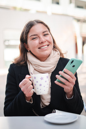 Joyful young woman enjoying a warm cup of coffee while using her smartphone to browse through applications in a delightful moment of relaxation andの写真素材