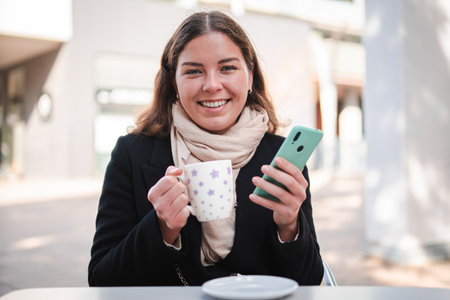 Smiling woman enjoying a warm cup of coffee while using her smartphone at a cafe table, engaged in browsing and connecting with friends through herの写真素材