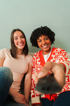 Two joyful women enjoying a fun movie night while excitedly using a remote control to change the channel and adjust the volume on their television forの写真素材