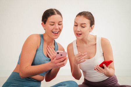 Two cheerful young women engaging with their smartphones while sharing joyful moments and laughter, exploring digital content and enjoying their timeの写真素材