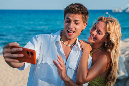 Happy couple taking a joyful selfie together on a sunny beach with a smartphone in their handsの写真素材